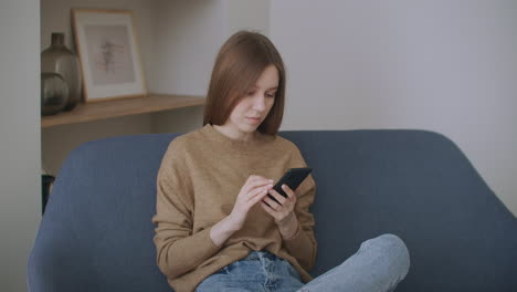 Una-Mujer-Escribiendo-Un-Mensaje-En-Su-Nuevo-Teléfono-Inteligente-Mientras-Está-Sentada-En-El-Sofá-Con-Una-Almohada-Blanca-En-Casa.